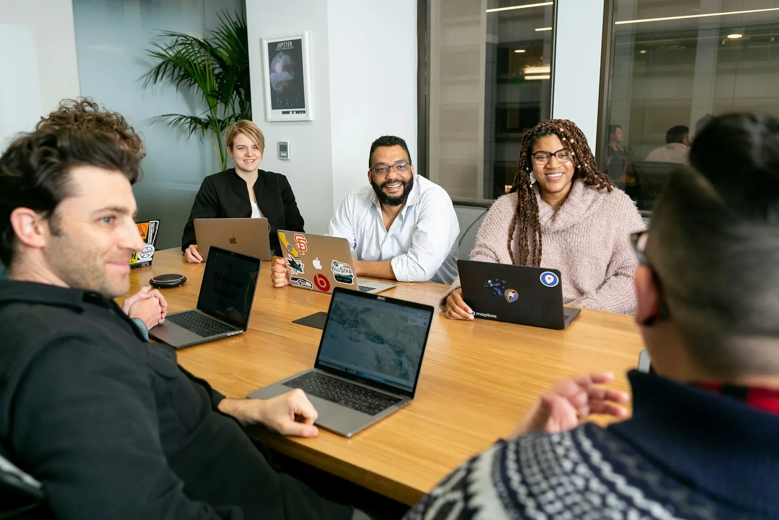 four people all on laptops, two men and two women, listen to person talking in a board meeting regarding benfit plans